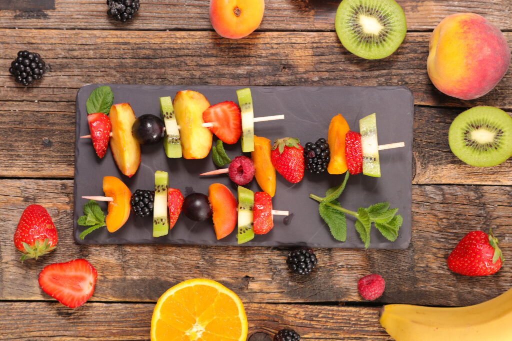 A serving plate with fruit skewers on a wooden table. The fruits include kiwi, strawberries, peaches, raspberries and blackberries. Garnished with mint leaves. 