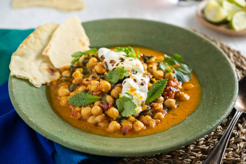 Healthy Eating Budget Recipe: The image shows a green stoneware bowl with chickpea and rainbow chard stew. The bowl is garnished with Greek yogurt and fresh mint leaves. It is served alongside two pieces of flame-baked naan.