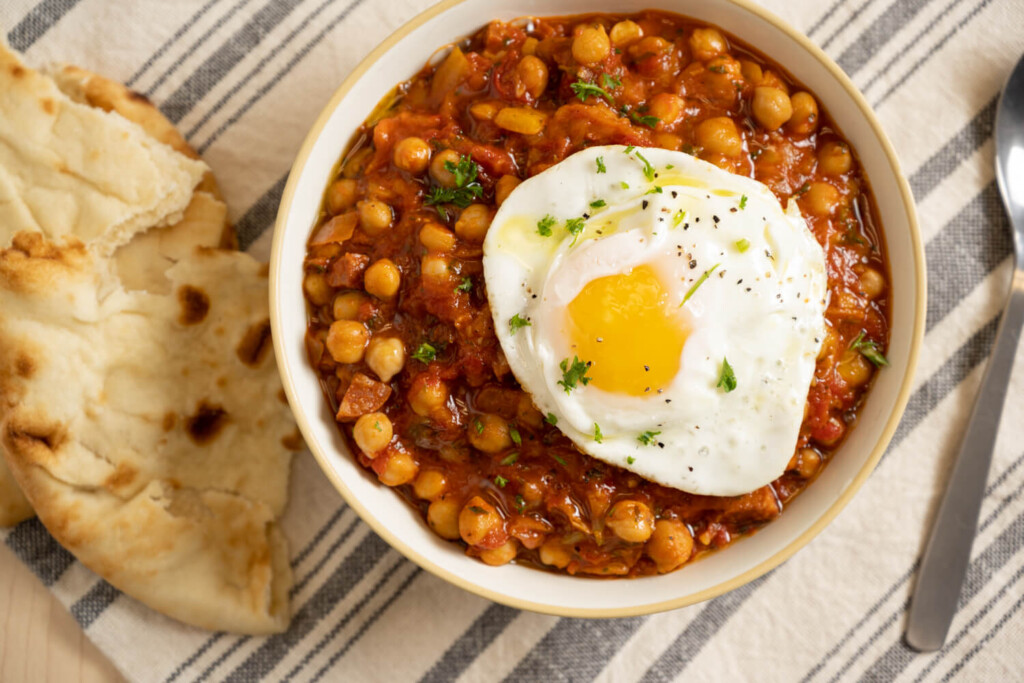 Overhead image of bowl of chickpea chorizo stew with fried egg on top beside hunk of crusty bread