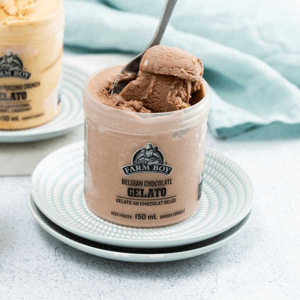 The image is taken from slightly above and shows two small pale blue plates on a pale countertop. On the plates is a single-serving container of Farm Boy Belgian Chocolate Gelato. Some of the gelato is being scooped out by a spoon. In the background out of focus on the right is a pale blue cloth. On the left out of focus in the background is a container of Farm Boy Toffee Cappuccino Crunch Gelato.