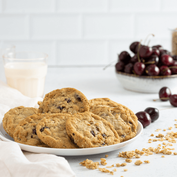 The image shows Plant-Based Cherry Granola Cookies fanned out on a white plate on a white countertop. In the background on the left side is a small glass of milk. On the right side in the background is a stoneware bowl filled with dark red sweet cherries. Four cherries have spilled out of the bowl. There is a trail of granola in the foreground of the image, beside the plate.