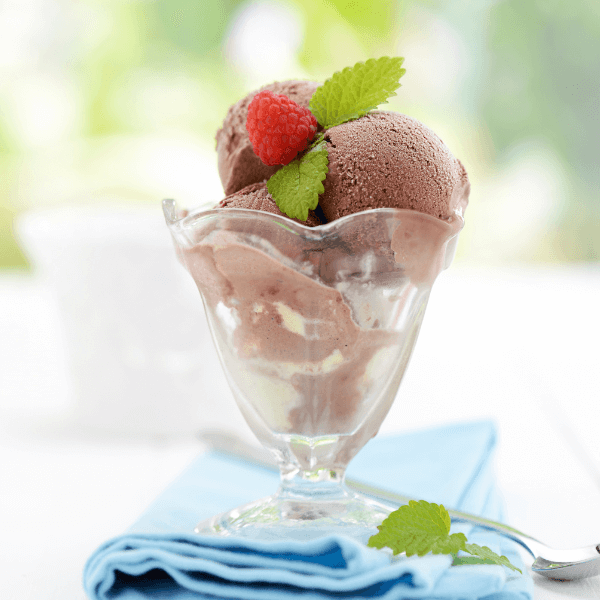 The image is taken outside and shows a parfait glass resting on a folded sky blue napkin. There is a spoon crossing the napkin diagonally. In the glass are scoops of chocolate ice cream. The ice cream is garnished with a fresh raspberry and two mint leaves.