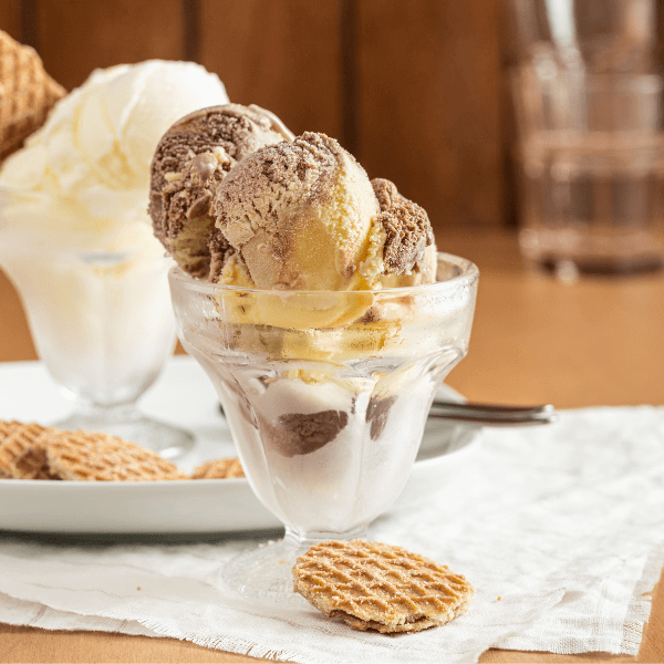The image shows a parfait glass on a piece of white linen. In the glass are scoops of Farm Boy Caramel Chocolate Swirl Ice Cream. At the foot of the glass is one Farm Boy Mini Stroopwafel. In the background, slightly out of focus on the left side is a matching parfait glass with scoops of Farm Boy Vanilla Ice Cream. The glass is on a white plate with more mini stroopwafel cookies at the foot.