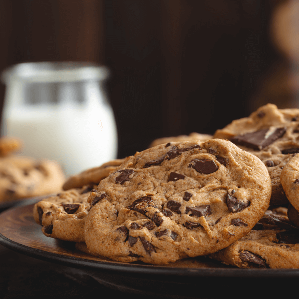 The image shows a close up of a plate with chocolate chunk cookies. The cookies look rich and chewy. In the background, out of focus, is a rounded glass of milk.