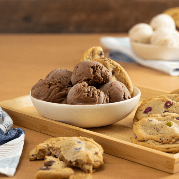 The image shows a wooden serving tray with a white bowl filled with scoops of Farm Boy Double Chocolate oat-rageous Frozen Dessert. there is a plant based monster cookie perched atop the scoops. Beside the bowl are two more soft-baked monster cookies. In the foreground is a monster cookie torn in half. Beside it, slightly out of frame in the lower left corner is a blue and white striped dishcloth. In the background upper right corner is another white bowl with scoops of vanilla ice cream.