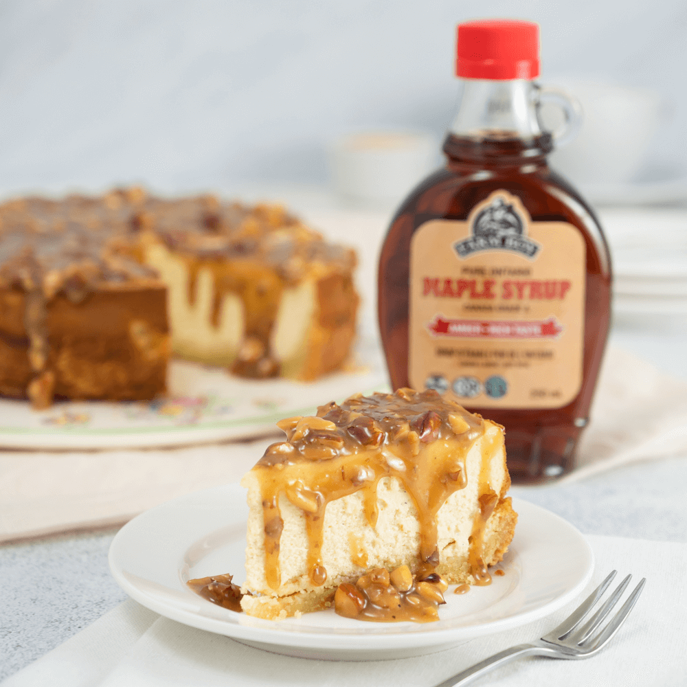The images shows a maple almond cheesecake with one slice removed. The slice is on a plate in the foreground. A bottle of Farm Boy Amber Maple Syrup in in the background.