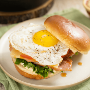 The image shows a stoneware plate with a breakfast sandwich. The breakfast sandwich is made of a brioche burger bun, lettuce, tomato, maple bacon, and a runny sunny side-up egg.