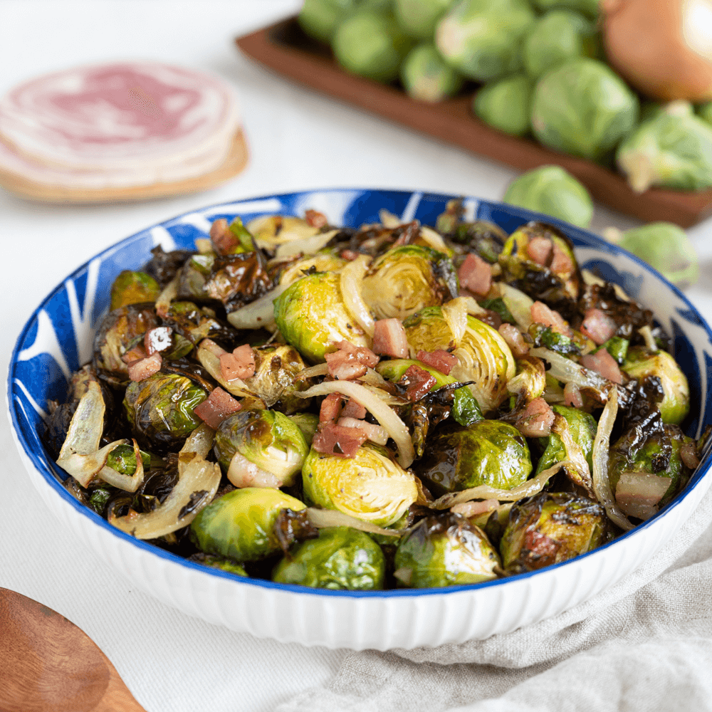 The image shows a blue and white bowl filled with roasted Brussels sprouts cooked with crispy pancetta and onions. Beside the bowl is a wooden spoon.