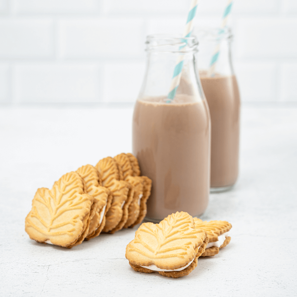 Two glass bottles of chocolate milk with straws behind six maple leaf-shaped maple-filled cream cookies.