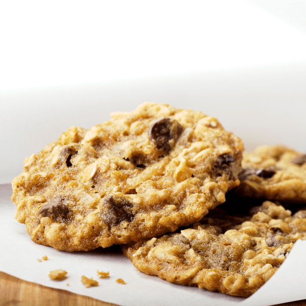 The image shows a close up of three oatmeal chocolate chip cookies. They are lying on a sheet of parchment paper on a wooden table. The cookies look soft and freshly baked.