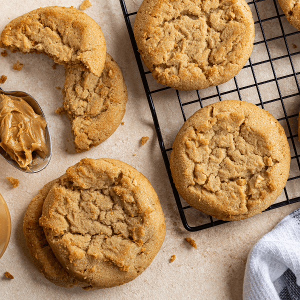 The image is taken from above and shows a countertop upon which peanut butter cookies are cooling. Two cookies are visible on a wire rack, while two other cookies are stacked towards the bottom of the image. Atop the stack is a cookie broken in half. At the left edge of the image a spoon with peanut butter is visible.