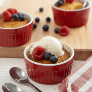Red ramekin filled with pouding chomeur topped with two blueberries, a raspberry, and a scoop of vanilla ice cream.