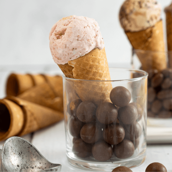 The image shows a sugar cone standing upright in a short glass tumbler. the tumbler is filled with round chocolate balls. In the cone is a scoop of Farm Boy Strawberry Marshmallow Twist Ice Cream. In the foreground of the image is a metal ice cream scoop. In the backgroundon the left side are more sugar cones. In the background on the left side is a similar glass, but it's filled with chocolate almonds. Two filled cones rest in this glass, but only the scoop of ice cream in one is visible.