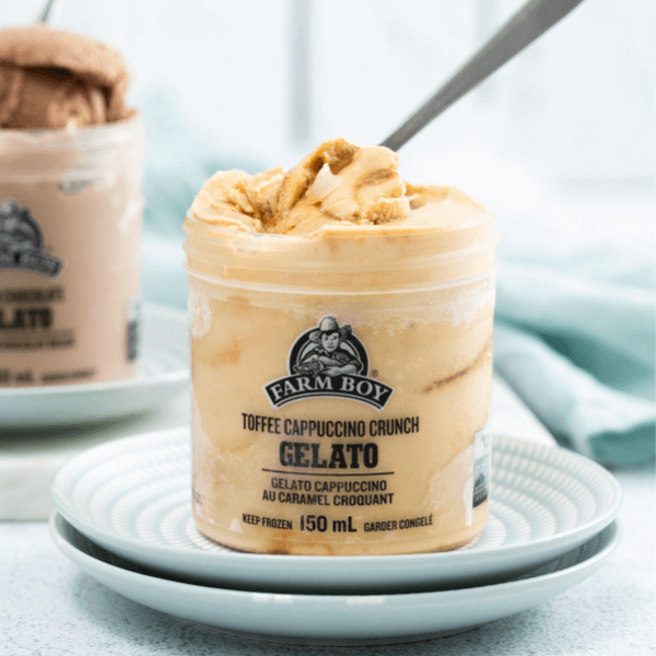The image shows two pale blue plates stacked on top of each other with a single-serve container of Farm Boy Toffee Cappuccino Crunch Gelato. Some of the gelato is being scooped out my a spoon. In the background out of focus on the left side is a single serve container of Farm Boy Belgian Chocolate Gelato. In the background out of focus is a pale blue cloth.