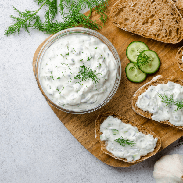 Overhead image of bowl of fresh tzatziki on wooden board with three slices of bread, two spread with tzatziki and garnished with fresh dill.