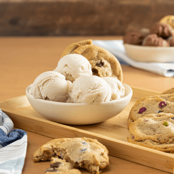 The image shows a wooden serving tray with a white bowl filled with scoops of Farm Boy Vanilla Bean oat-rageous Frozen Dessert. there is a plant based monster cookie perched atop the scoops. Beside the bowl are two more soft-baked monster cookies. In the foreground is a monster cookie torn in half. Beside it, slightly out of frame in the lower left corner is a blue and white striped dishcloth. In the background upper right corner is another white bowl with scoops of chocolate ice cream.