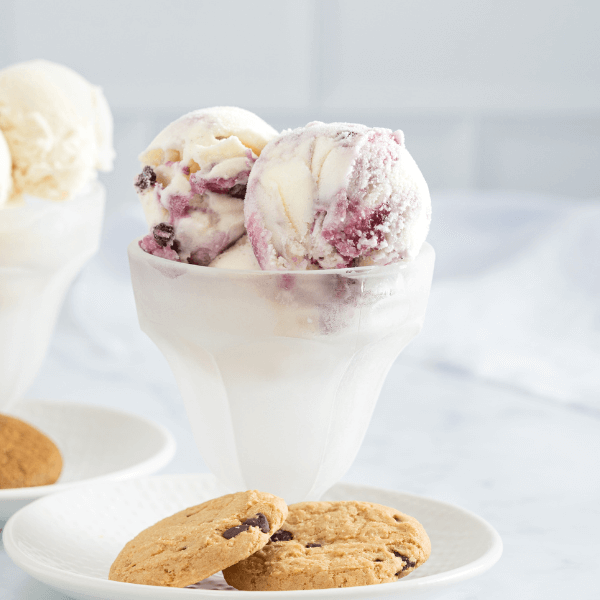 The image shows a frosty parfait glass on a small white plate. In the glass are two visible scoops of Farm Boy Wild Blueberry Boom Ice Cream. At the foot of the glass are two chocolate chip cookie chips. In the background out of focus on the left is a matching parfait glass on a white plate, but this glass is filled with scoops of vanilla ice cream.