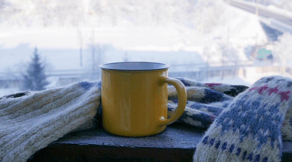 Yellow cup with hot tea wrapped with a woolen scarf on a wooden surface against the backdrop of a snowy landscape, cold season, winter, morning